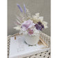 Dried & Preserved flower arrangement in purple & white and featuring a purple preserved rose and nestled in to a white pot. Photo shows arrangement sitting on a right angle view on a book on a table against a blank wall
