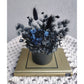 Dried & Preserved flower arrangement in black & navy blue colours and nestled in to a mini black pot. Photo shows arrangement sitting on a book against a blank wall