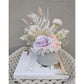 Dried & Preserved flower arrangement in pastel pink, purple & white flowers and set in to a light grey cement pot. Photo shows arrangement sitting on a book against a blank wall