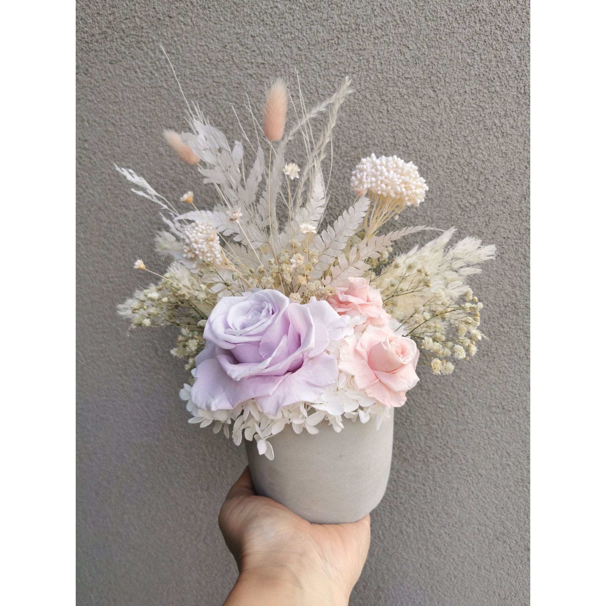 Dried & Preserved flower arrangement in pastel pink, purple & white flowers and set in to a light grey cement pot. Photo shows arrangement being held by hand against a blank wall