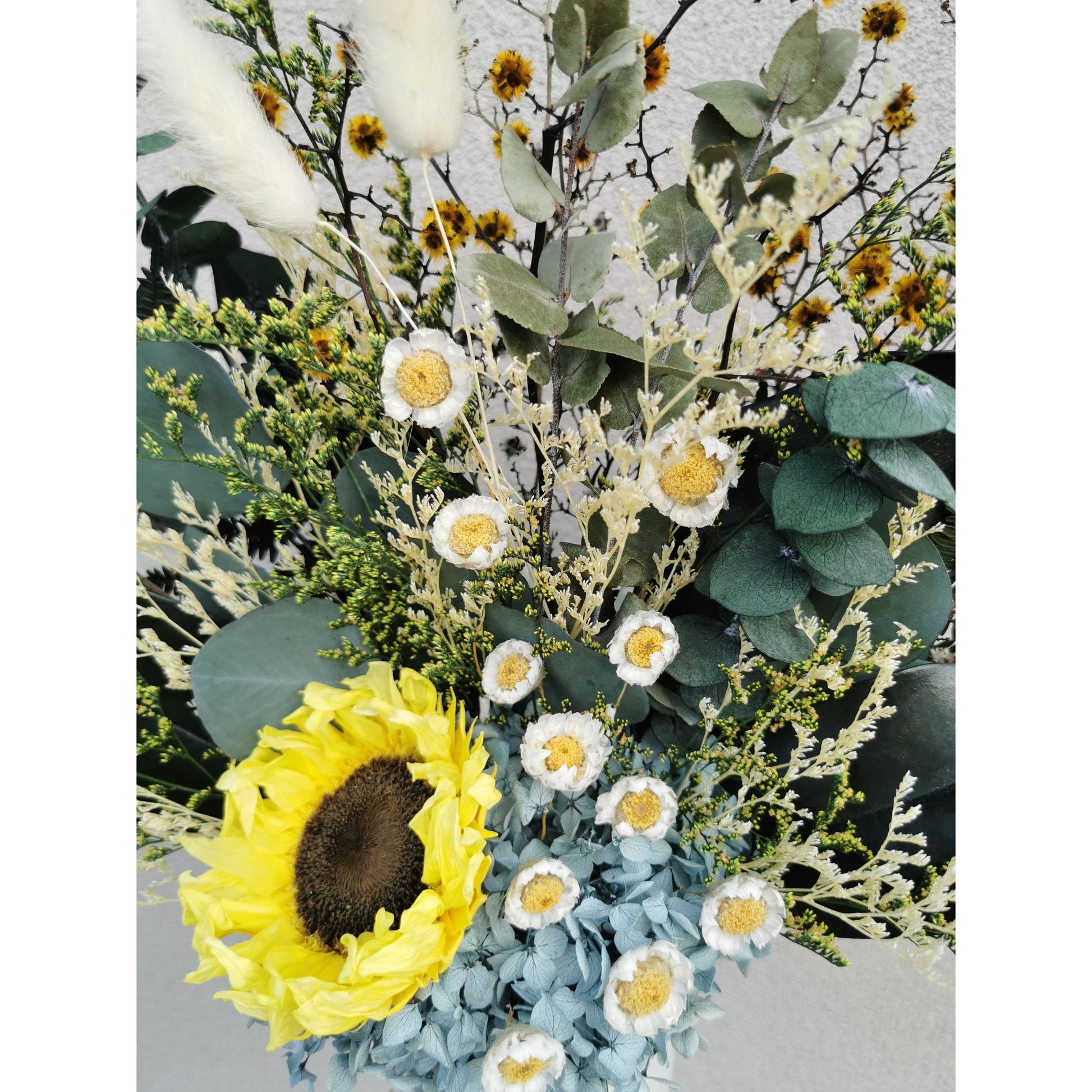 Dried & Preserved flower arrangement featuring a preserved yellow sunflower and yellow daisies and lush greenery and set in to a white pot. Photo shows a birds eye view of the flowers and the daisies cascading down the arrangement