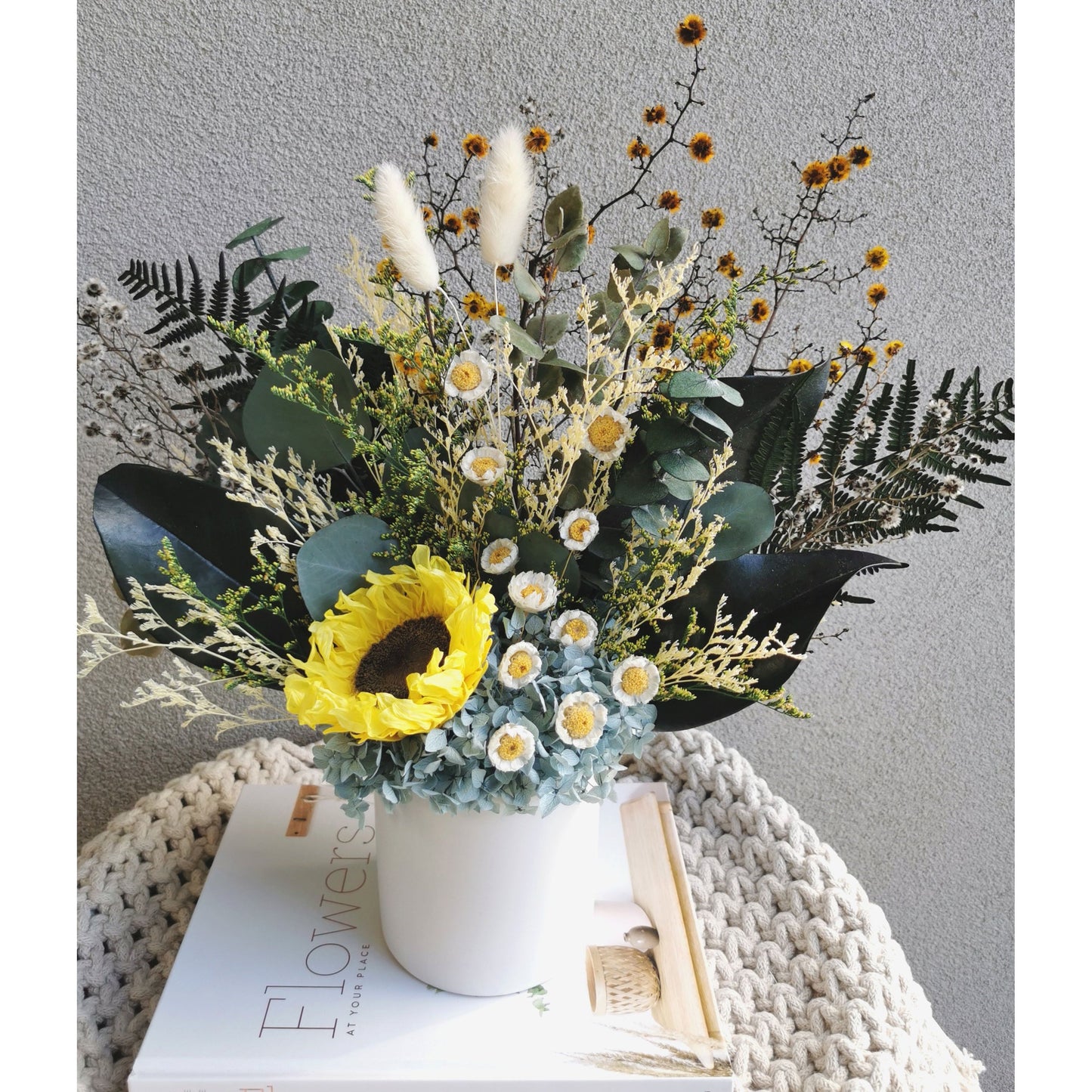Dried & Preserved flower arrangement featuring a preserved yellow sunflower and yellow daisies and lush greenery and set in to a white pot. Photo shows arrangement sitting on a book against a blank wall