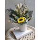Dried & Preserved flower arrangement featuring a preserved yellow sunflower and yellow daisies and lush greenery and set in to a white pot. Photo shows arrangement sitting on a book at an angle against a blank wall