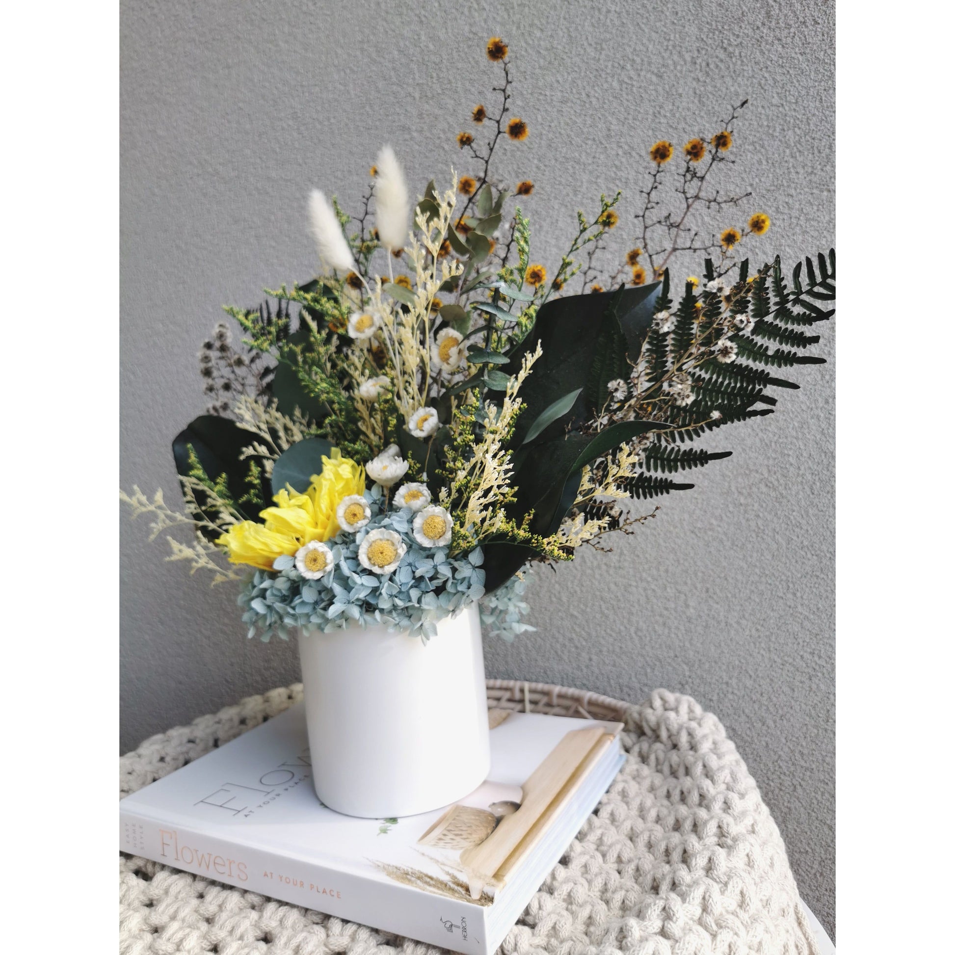 Dried & Preserved flower arrangement featuring a preserved yellow sunflower and yellow daisies and lush greenery and set in to a white pot. Photo shows arrangement sitting on a book on a side angle against a blank wall