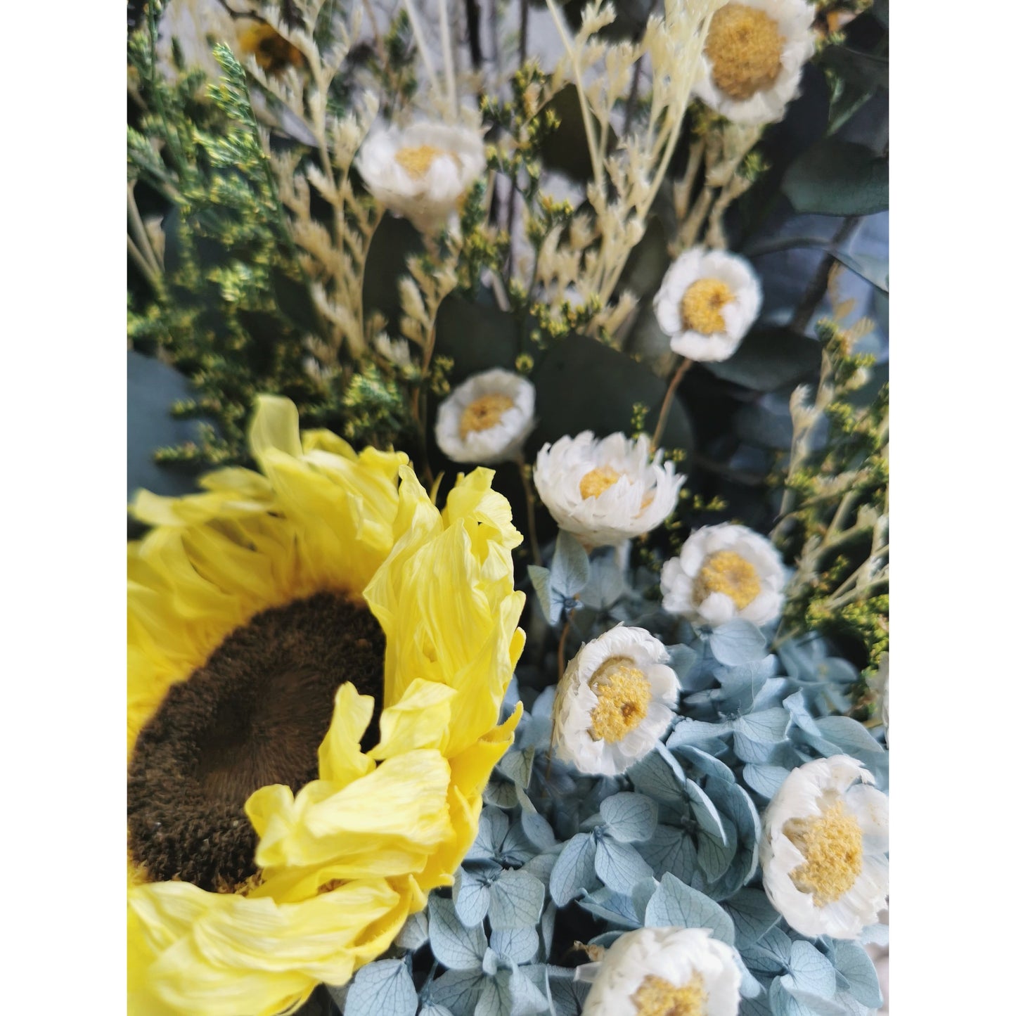 Dried & Preserved flower arrangement featuring a preserved yellow sunflower and yellow daisies and lush greenery and set in to a white pot. Photo shows a zoomed in up close photo of the flowers