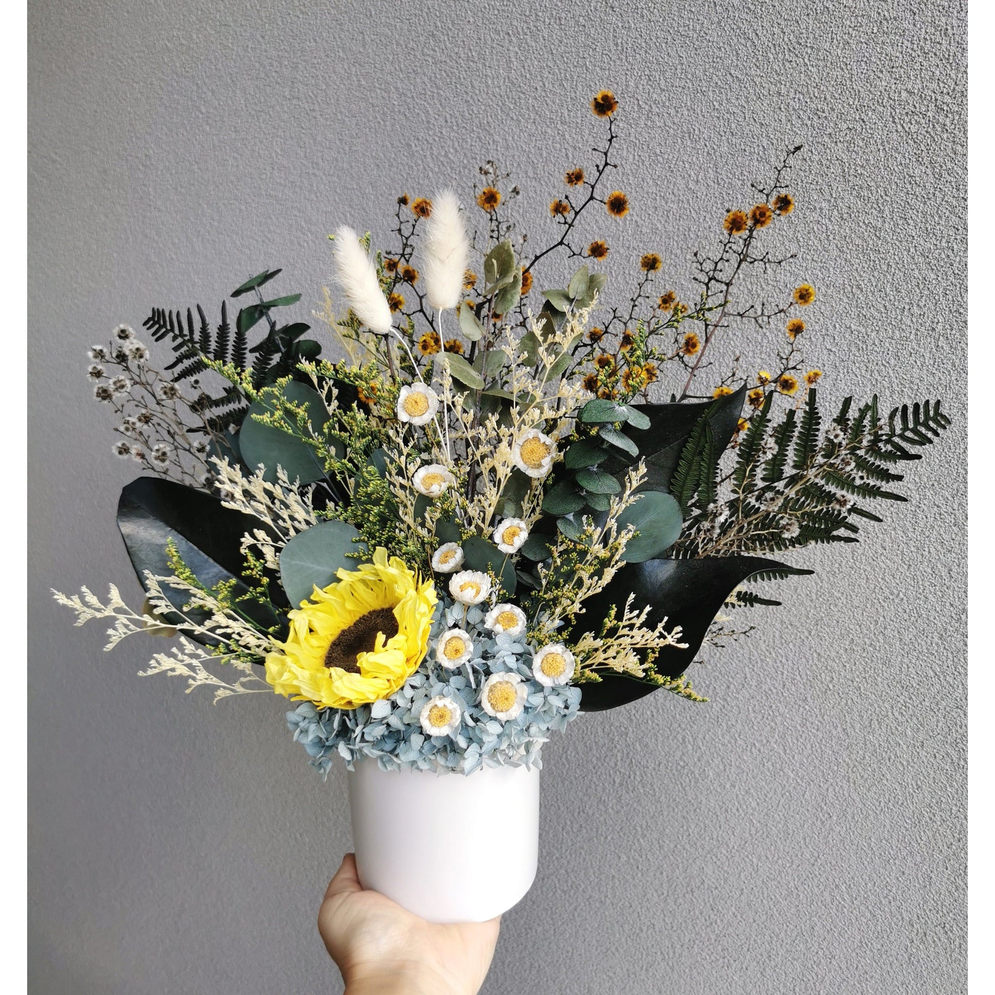 Dried & Preserved flower arrangement featuring a preserved yellow sunflower and yellow daisies and lush greenery and set in to a white pot. Photo shows arrangement being held by hand against a blank wall