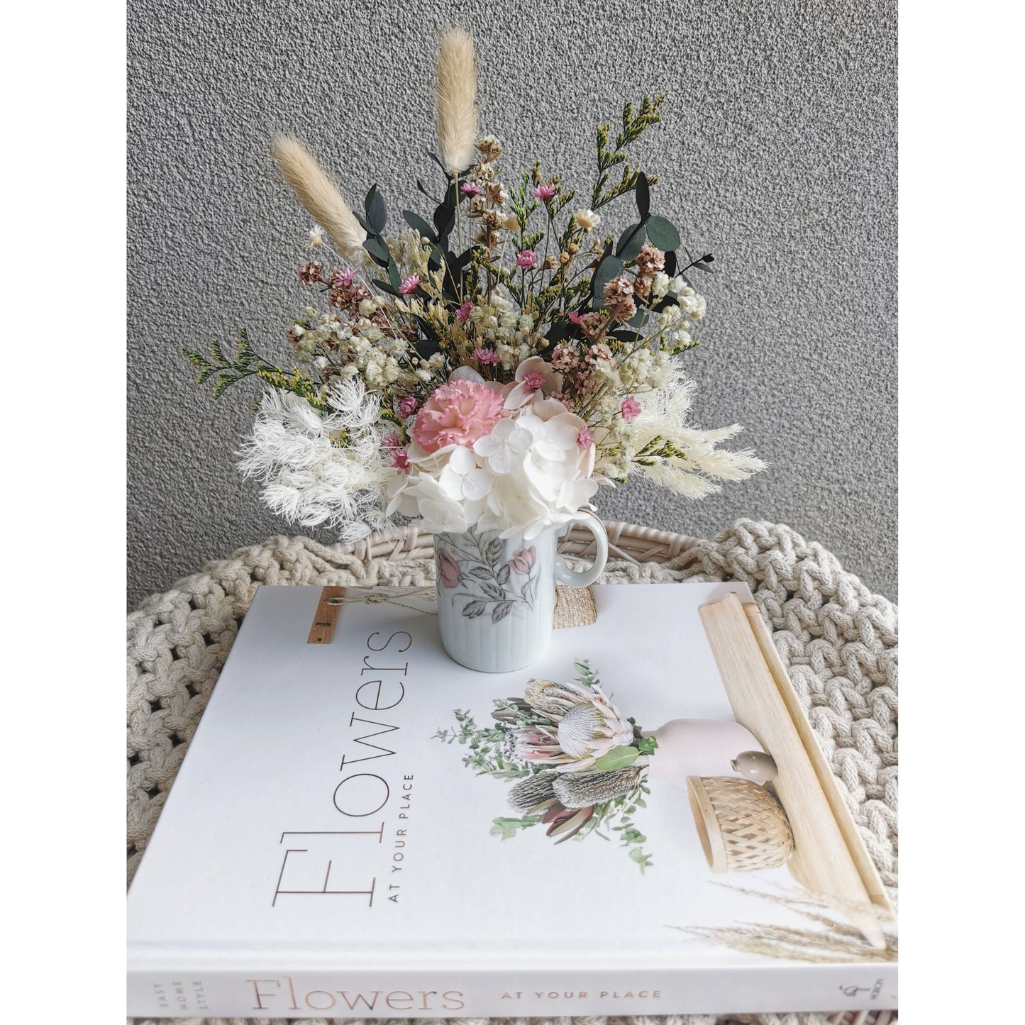 Mini Dried & Preserved flower arrangement with green, pink & white flowers in a mini geisha bone China Japan vase with handle. Photo shows arrangement sitting on a book against a blank wall