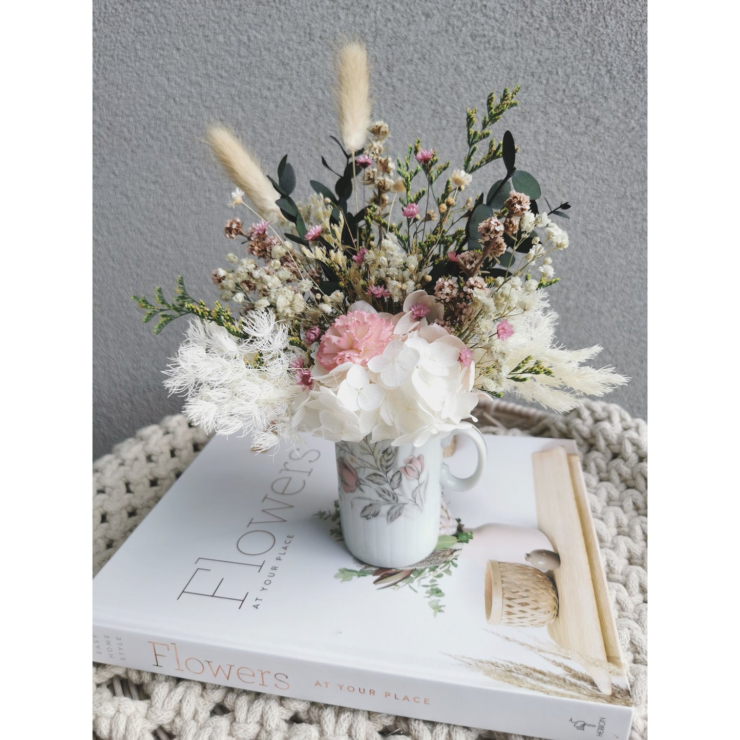 Mini Dried & Preserved flower arrangement with green, pink & white flowers in a mini geisha bone China Japan vase with handle. Photo shows arrangement sitting on a book on an angle against a blank wall