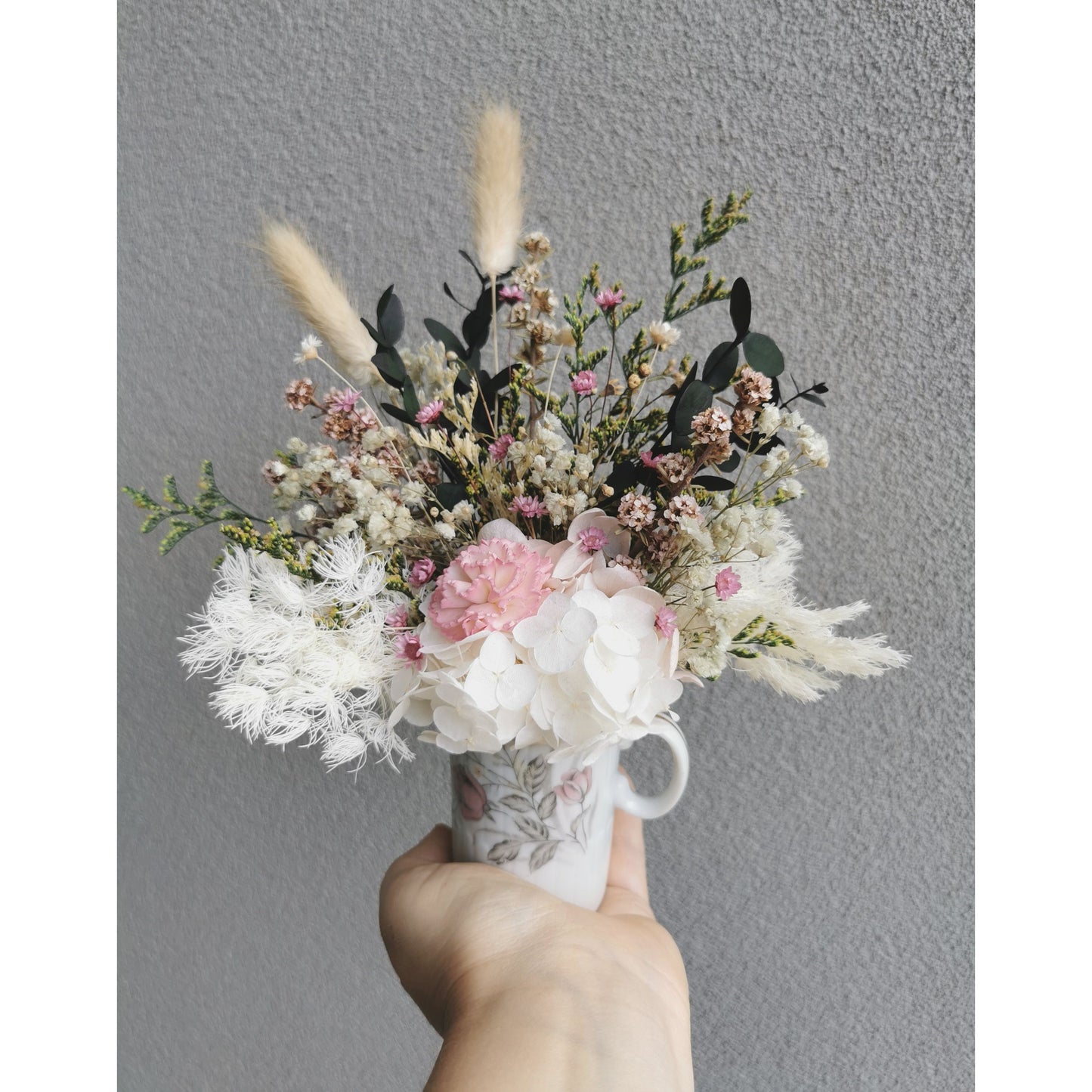 Mini Dried & Preserved flower arrangement with green, pink & white flowers in a mini geisha bone China Japan vase with handle. Photo shows arrangement being held by hand against a blank wall