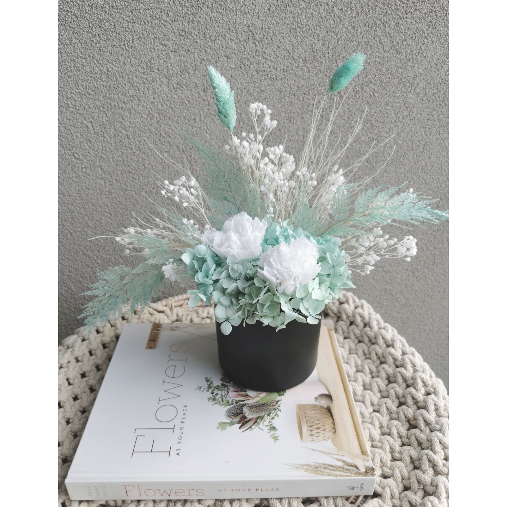 Dried & Preserved flower arrangement in Tiffany Blue & White colours and set in to a black pot. Photo shows arrangement sitting on a book against a blank wall