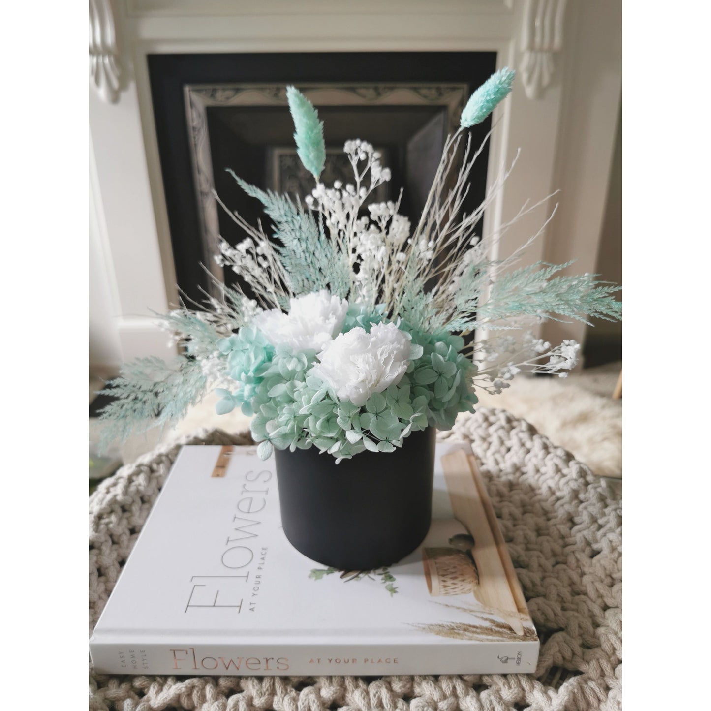 Dried & Preserved flower arrangement in Tiffany Blue & White colours and set in to a black pot. Photo shows arrangement sitting on a book on a coffee table with a fireplace in the background