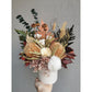 Dried & Preserved native flower arrangement in browns, greens, neutral colours and comes in a white pot including banksias, ferns & bunny tails to name a few. Photo shows arrangement being held by hand against a blank wall