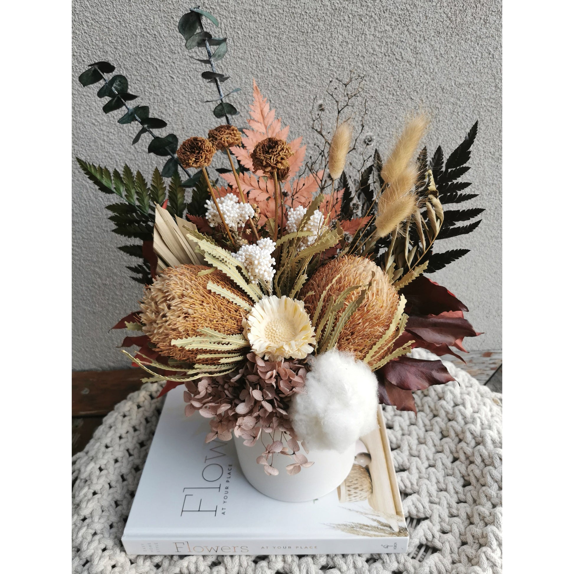 Dried & Preserved native flower arrangement in browns, greens, neutral colours and comes in a white pot including banksias, ferns & bunny tails to name a few. Photo shows arrangement sitting on a book against a blank wall