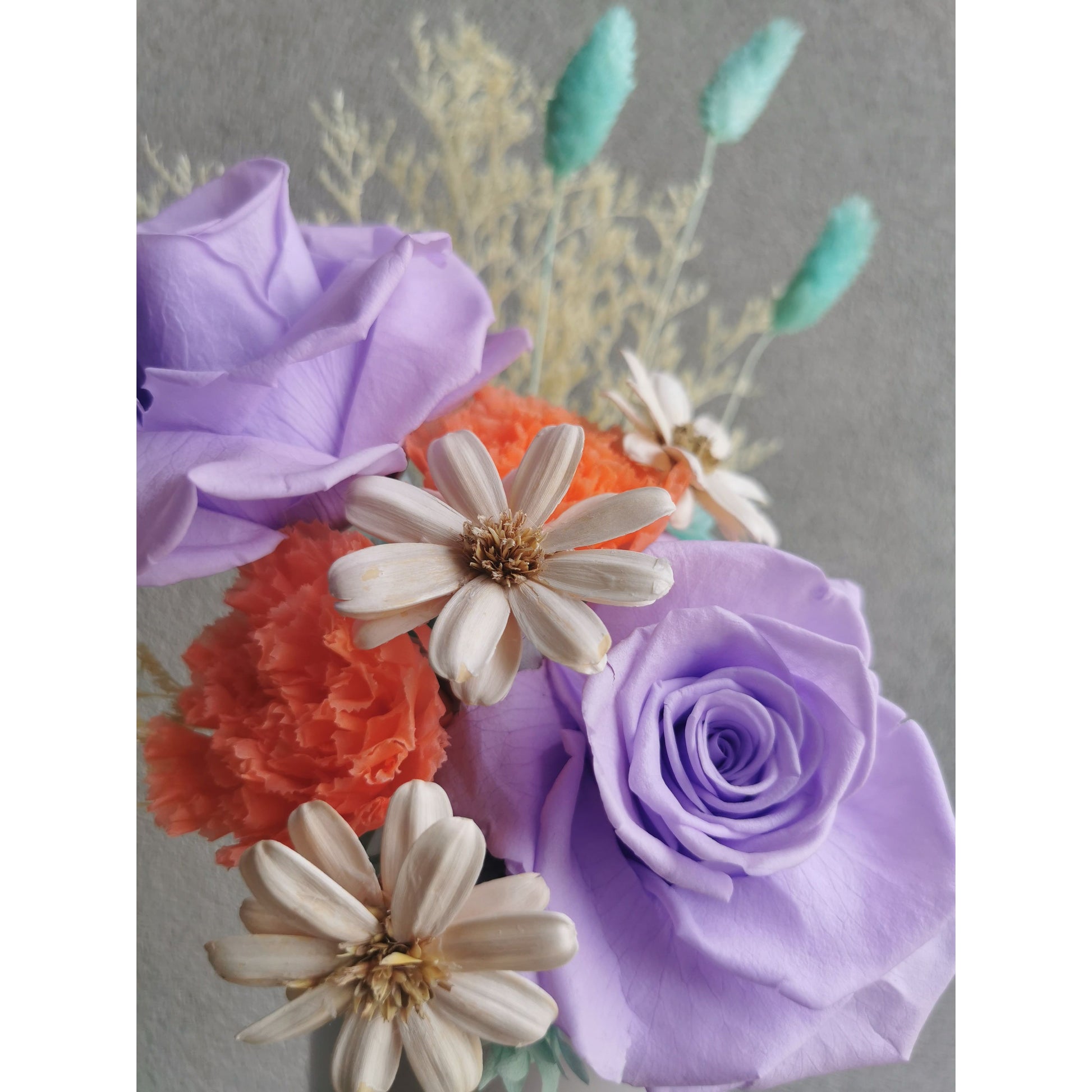Dried & Preserved flower arrangement in purple, orange, white, blue & yellow. Featuring purple preserved roses and orange preserved carnations. Flowers are set in to a white pot. Photo shows a close up photo of the flowers