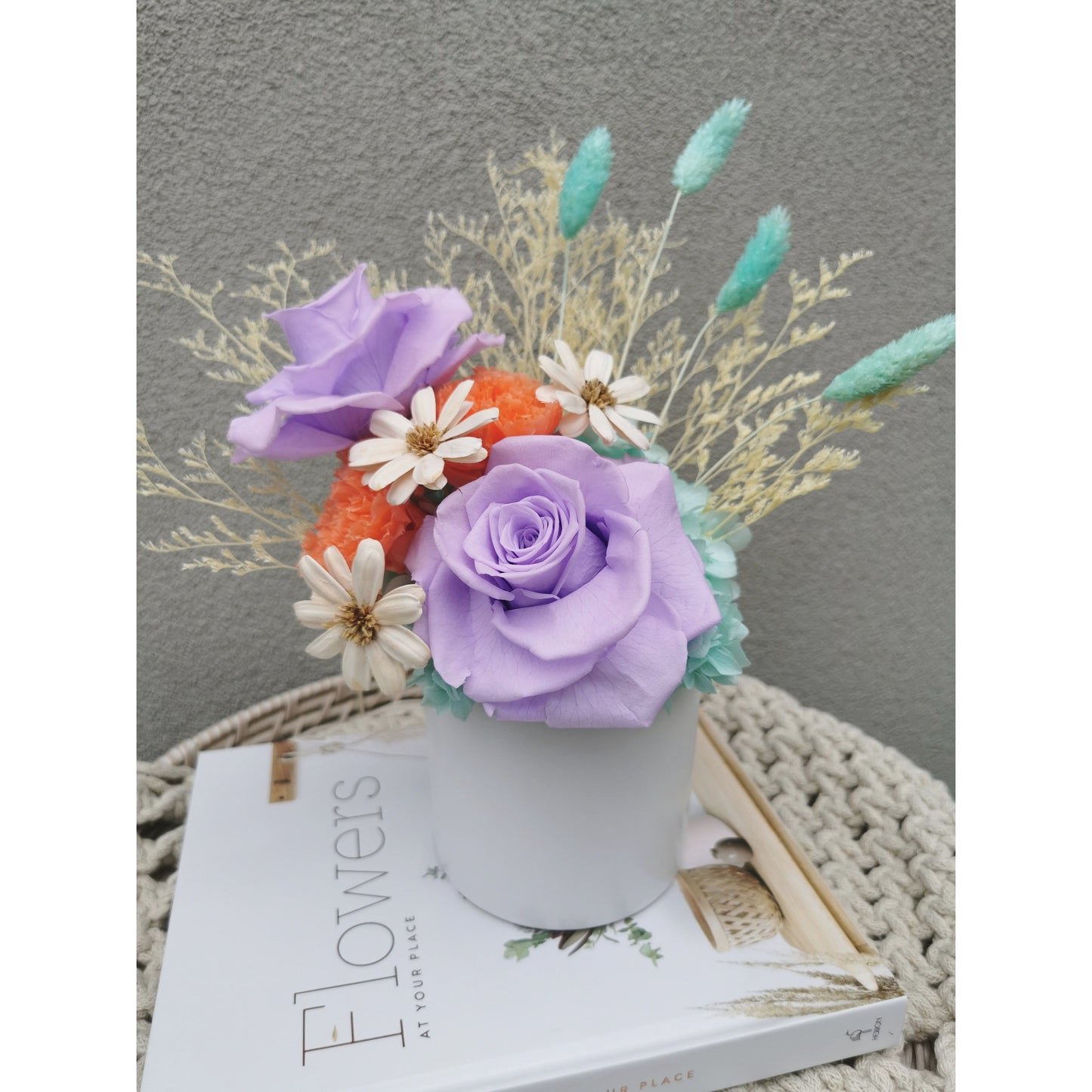 Dried & Preserved flower arrangement in purple, orange, white, blue & yellow. Featuring purple preserved roses and orange preserved carnations. Flowers are set in to a white pot. Photo shows arrangement sitting on a book on an angle against a blank wall
