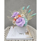 Dried & Preserved flower arrangement in purple, orange, white, blue & yellow. Featuring purple preserved roses and orange preserved carnations. Flowers are set in to a white pot. Photo shows arrangement sitting on a book against a blank wall