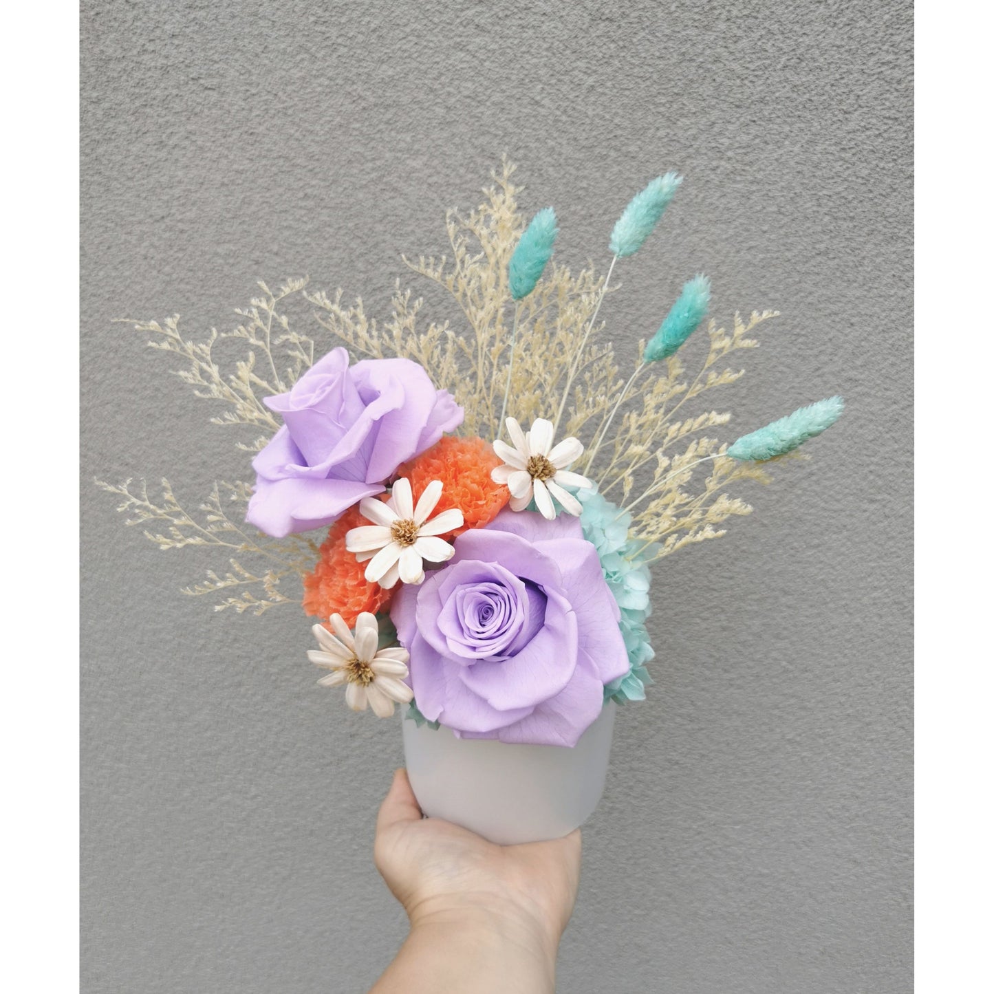 Dried & Preserved flower arrangement in purple, orange, white, blue & yellow. Featuring purple preserved roses and orange preserved carnations. Flowers are set in to a white pot. Photo shows arrangement being held by hand against a blank wall