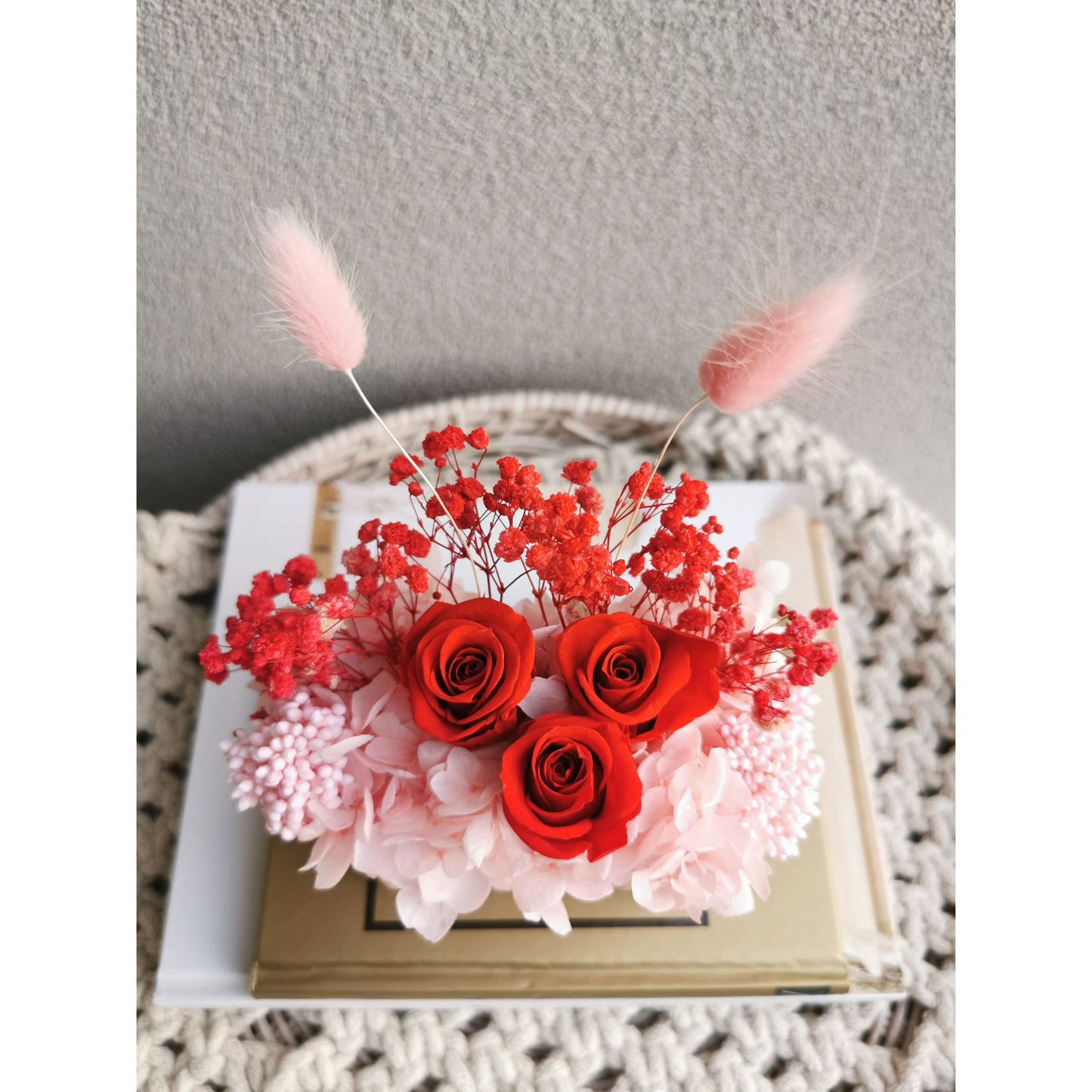 Valentines Day dried & preserved flower arrangement with red & pink flowers in pink arch vase. Photo shows arrangement sitting on books against a blank wall from a birds eye view of the flowers