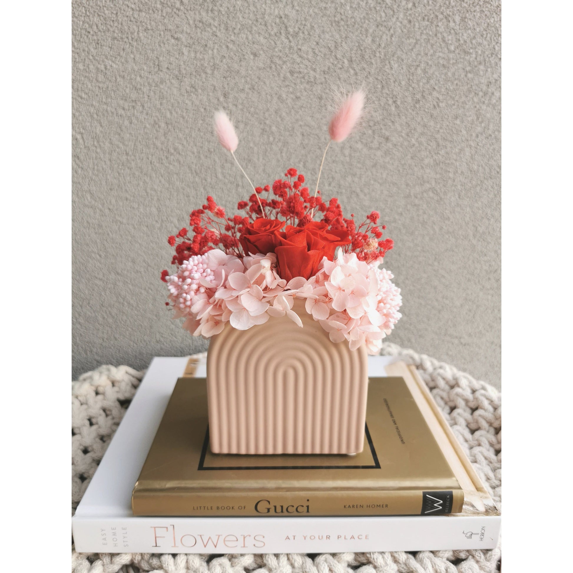 Valentines Day dried & preserved flower arrangement with red & pink flowers in pink arch vase. Photo shows arrangement sitting front on, on books against a blank wall