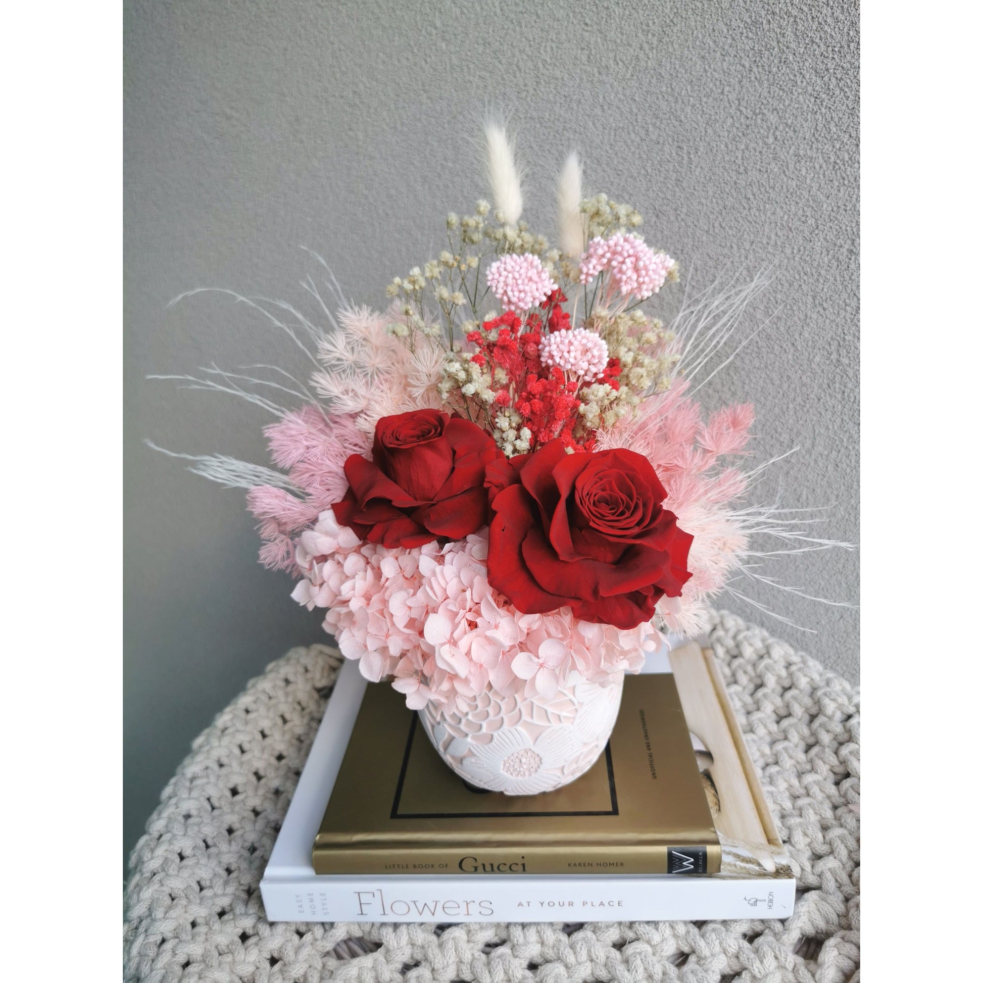 Valentines Day dried & preserved flower arrangement including 2 red roses and set in to a pink & white floral pot. Photo shows arrangement sitting on books against a blank wall