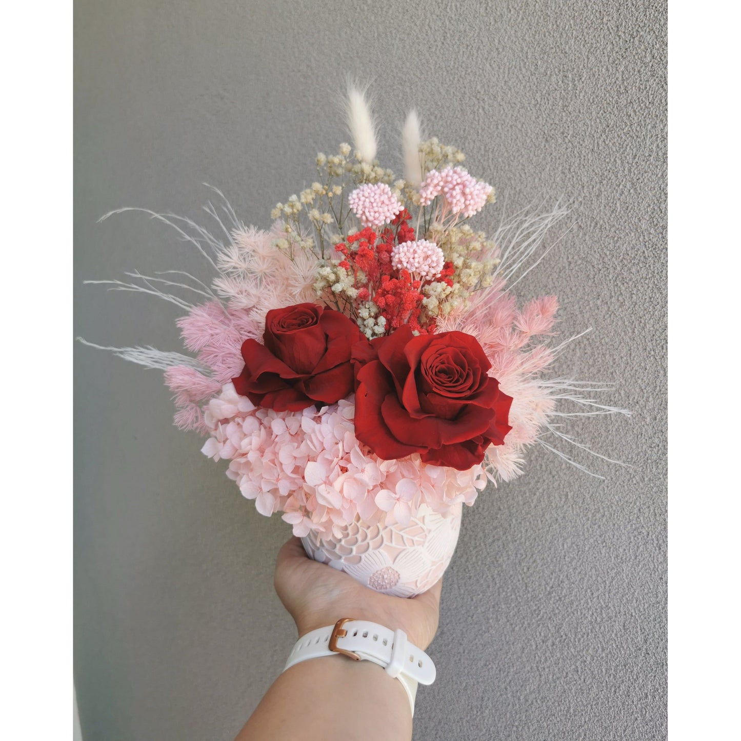Valentines Day dried & preserved flower arrangement including 2 red roses and set in to a pink & white floral pot. Photo shows arrangement being held by hand against a blank wall