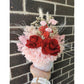 Valentines Day dried & preserved flower arrangement including 2 red roses and set in to a pink & white floral pot. Photo shows arrangement being held by hand against a black brick wall