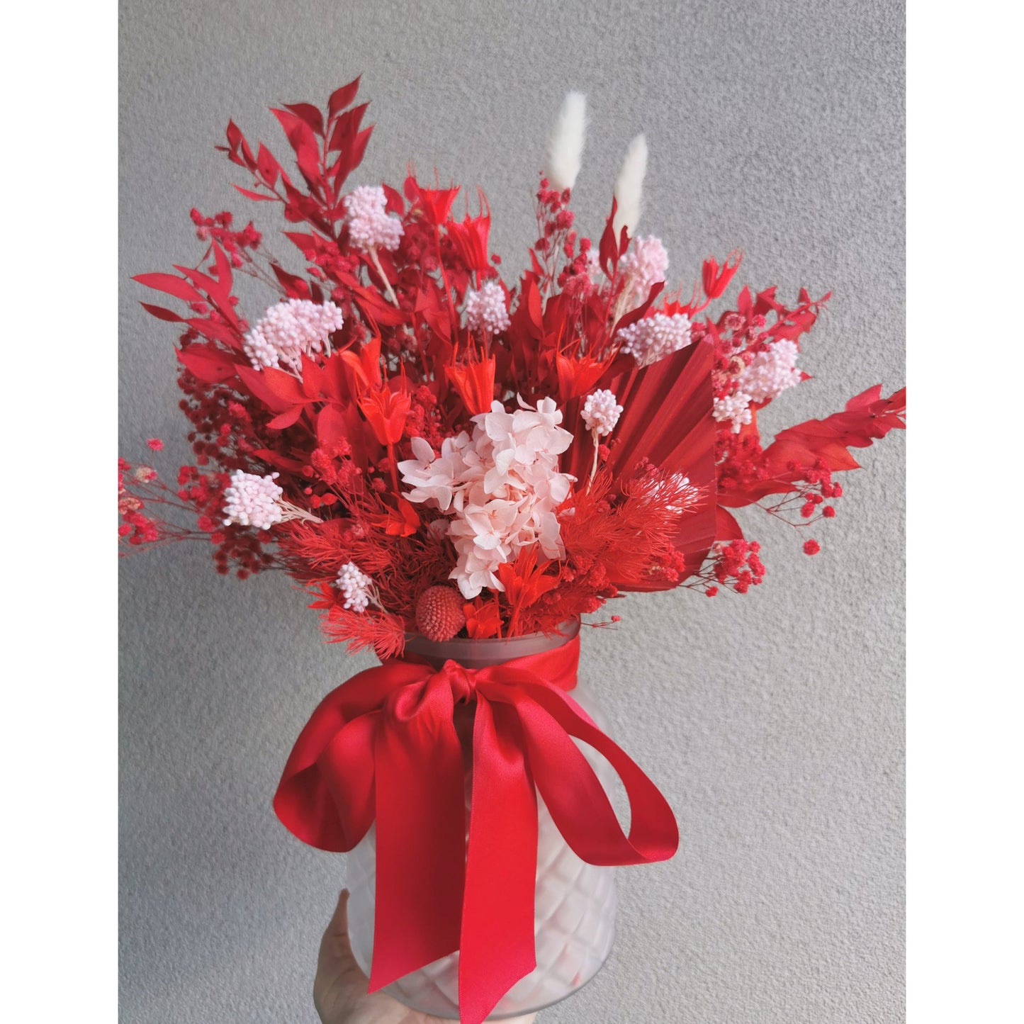 Bunch of Dried & Preserved flowers in red & pink colours and sitting in a frosted glass vase. Photo shows arrangement being held by hand against a blank wall